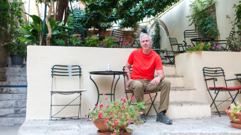 Seated man in a red shirt at a small courtyard café with metal chairs, stone steps, greenery, and flowering pots.