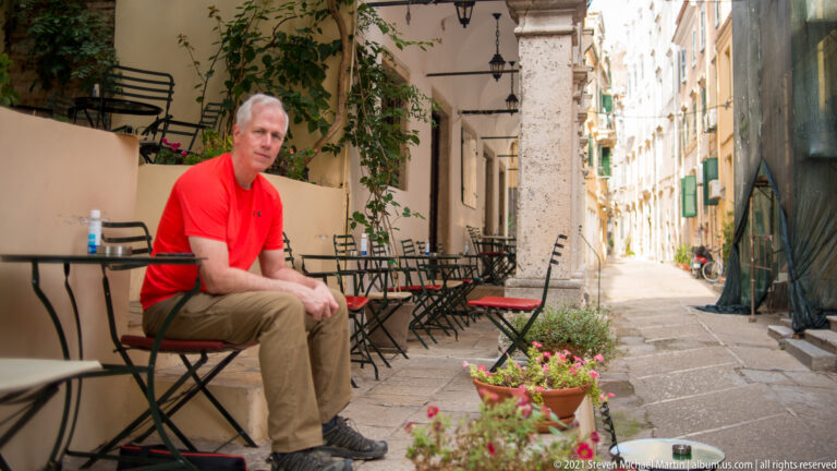 Man in red shirt seated beside café tables under an arcade, with a narrow sunlit Corfu lane extending behind him.