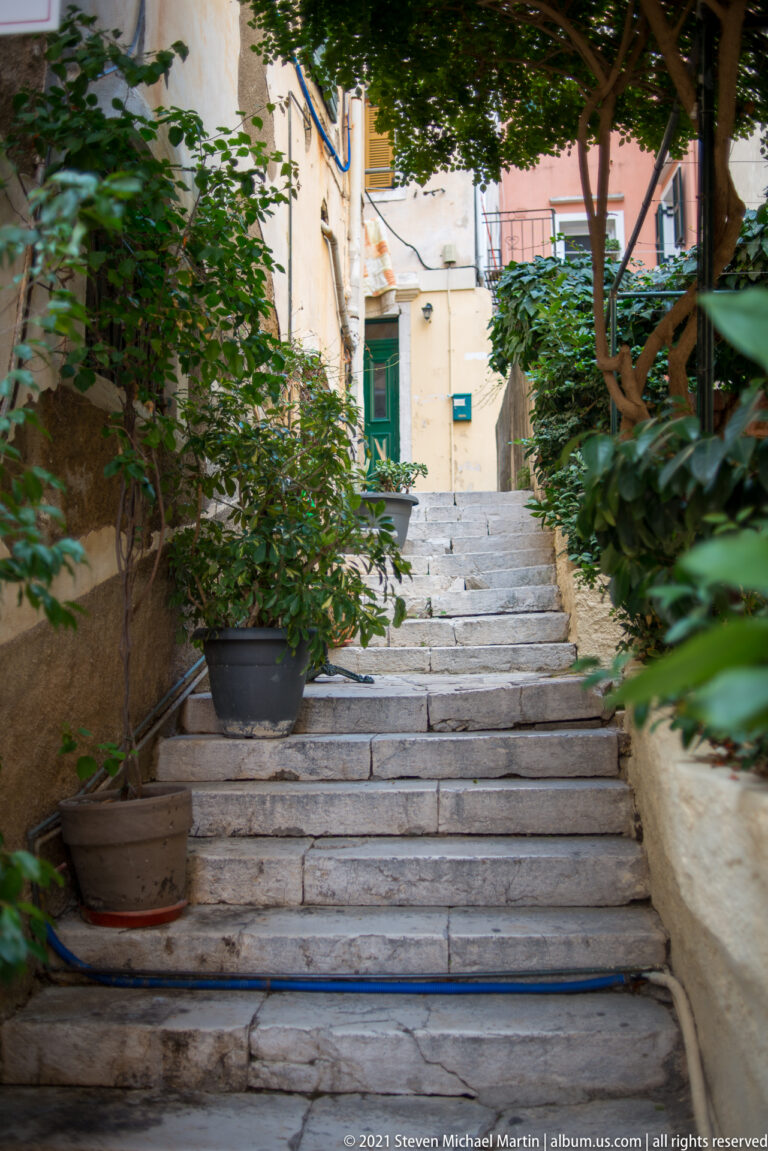Wide stone steps rising through a leafy Corfu passage toward pale buildings with green shutters.