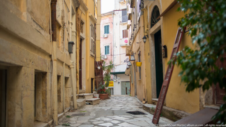 Narrow Corfu alley with stone paving, ocher walls, shutters, wall lamps, potted plants, and closely spaced buildings.