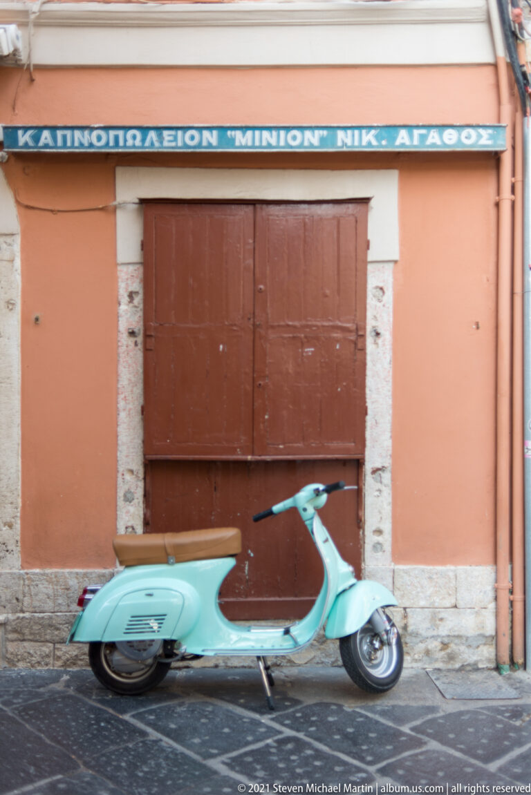 Turquoise vintage-style step-through scooter parked against a salmon wall beneath a closed brown shop shutter.