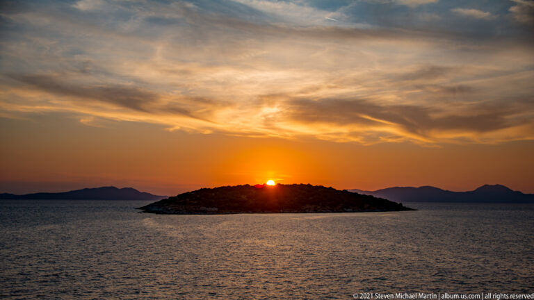 Sun setting directly behind a low dark island, with calm sea and distant landforms under streaked clouds.
