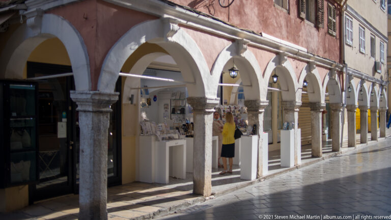 Long Corfu arcade with pale stone arches and columns, hanging lanterns, shops, and displays beneath a weathered upper façade.