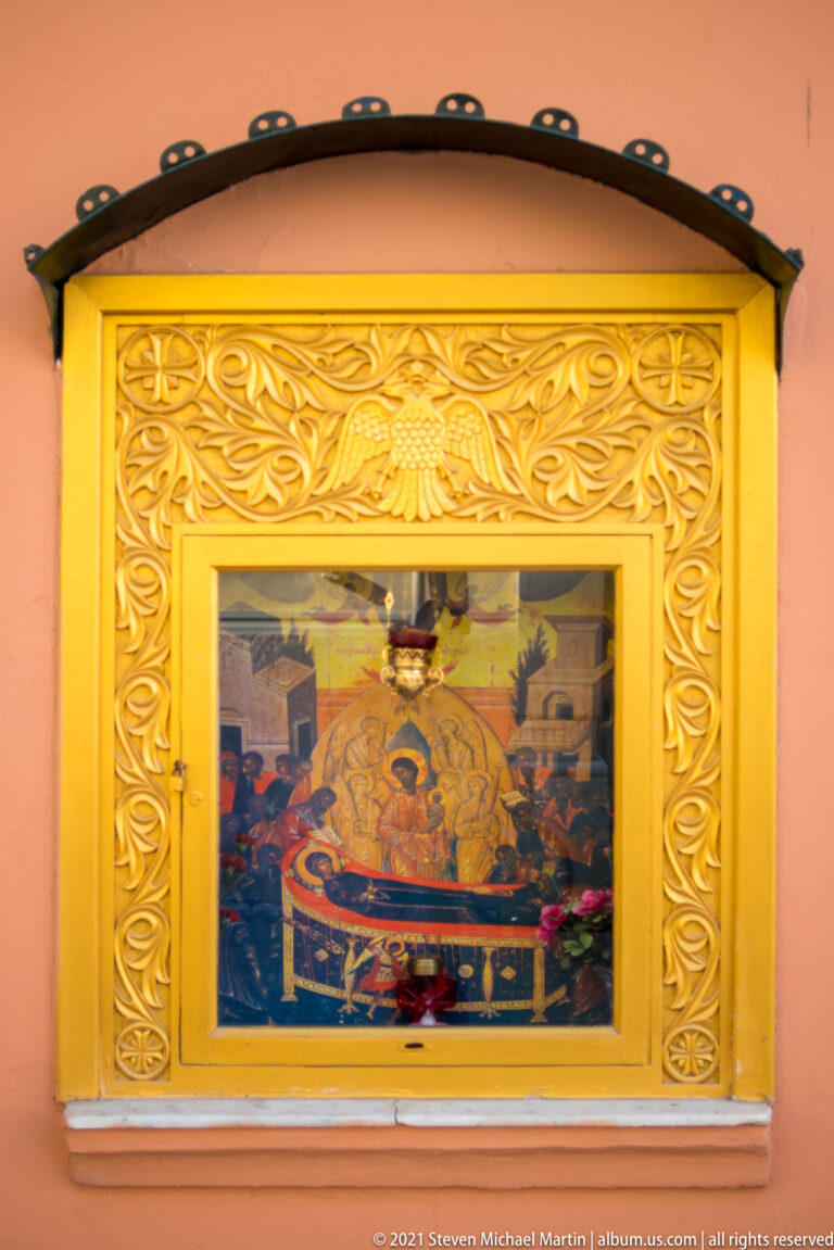 Orthodox devotional image behind glass in an ornate gold frame, with a small red votive lamp on a salmon wall.