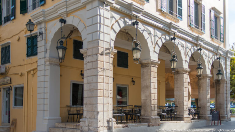 Rusticated pale-stone arcade with tall round arches, black hanging lanterns, green shutters, and café seating.