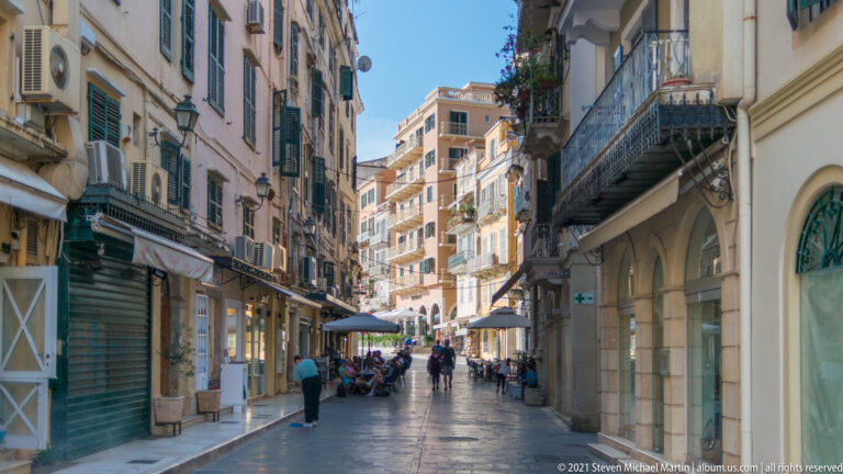 Busy paved Corfu shopping street with pedestrians, cafés, awnings, balconies, shutters, and tall historic buildings.