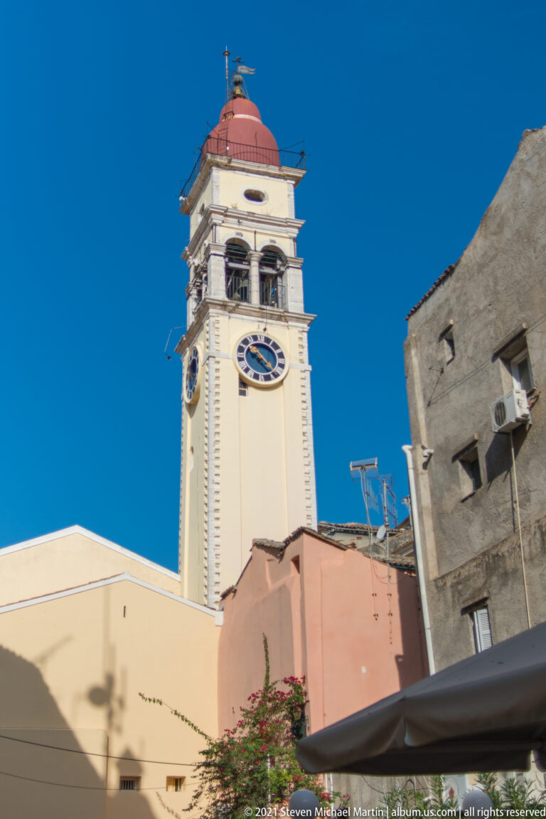Low-angle view of a cream clock tower with large clock faces, open belfry, and red domed roof above nearby buildings.