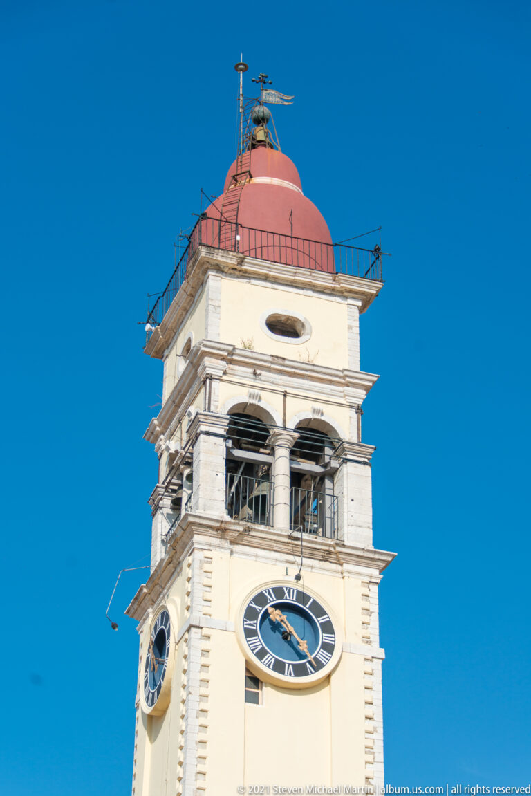 Close low-angle view of a cream clock tower with Roman-numeral clock face, open belfry, red dome, and rooftop railing.