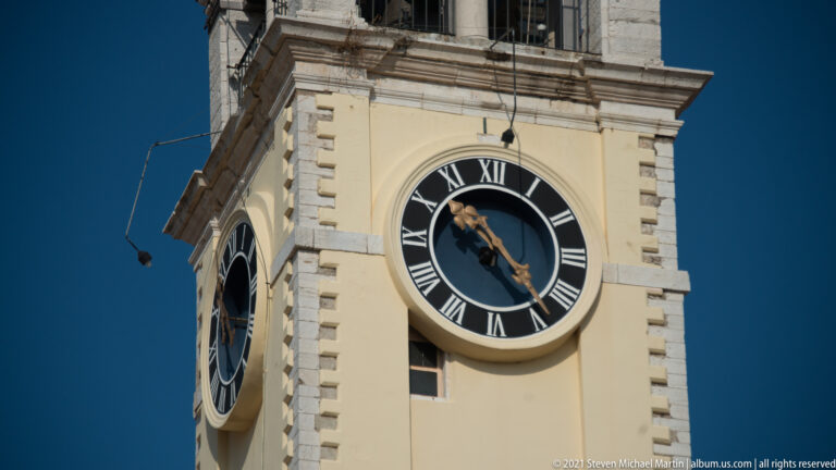 Large black clock face with white Roman numerals and ornate hands on a cream tower, with a second dial visible around the corner.