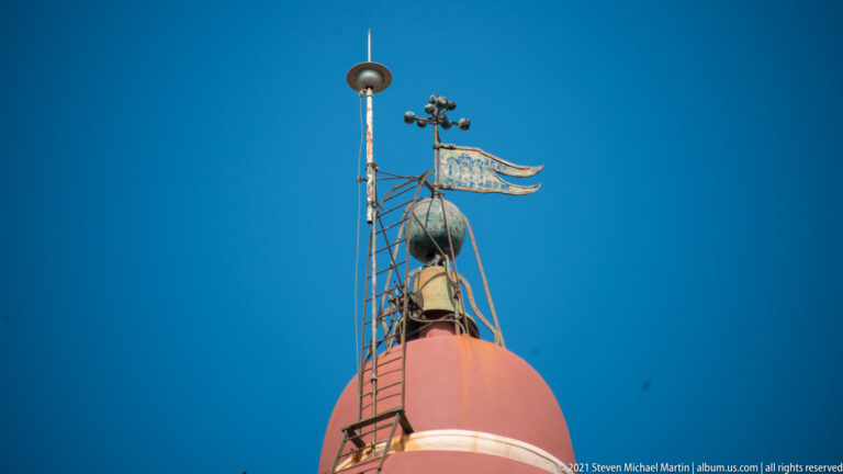 Close view of a red clock-tower dome topped by metal ladders, railings, a small bell-like element, and weather apparatus.