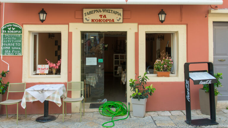 Small salmon-colored Corfu taverna with Greek sign, open doorway, sidewalk table and chairs, potted plants, and green hose.