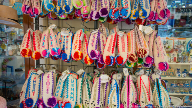 Rows of colorful embroidered slippers with braided trim and pom-poms hanging outside a Corfu souvenir shop.