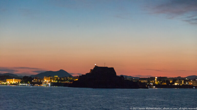 Old Fortress of Corfu silhouetted above the water at dusk, with waterfront lights and a fading pastel sky.