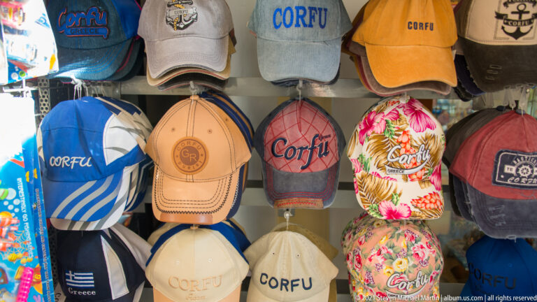 Souvenir baseball caps in varied colors and floral patterns marked Corfu or Greece and displayed in dense rows.
