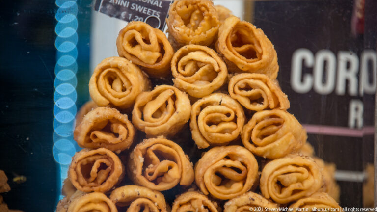 Stack of golden spiral fried pastries with crisp bubbled edges and syrupy surfaces in a Corfu confectionery display.
