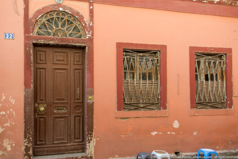 Weathered salmon façade with dark paneled wooden door, two metal-barred windows, red-brown stone trim, and peeling plaster.