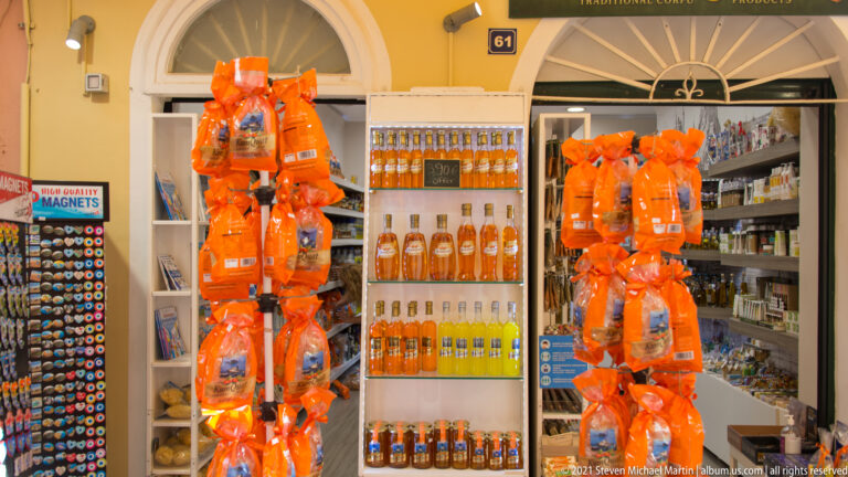 Corfu souvenir storefront with orange gift bags, rows of amber and yellow bottled products, magnets, and other packaged goods.