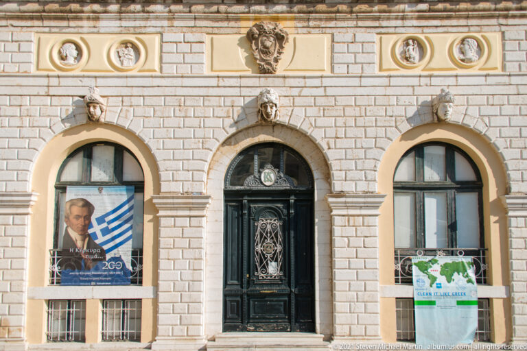 Stone-faced Corfu building with black arched central door, matching arched windows, sculpted heads, relief medallions, and posters.
