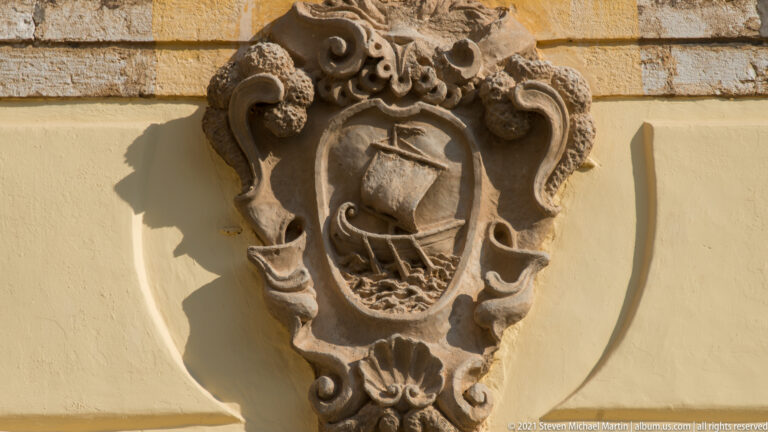 Weathered stone cartouche with a shield carved with a sailing vessel and waves, surrounded by scrolling ornament on a yellow wall.