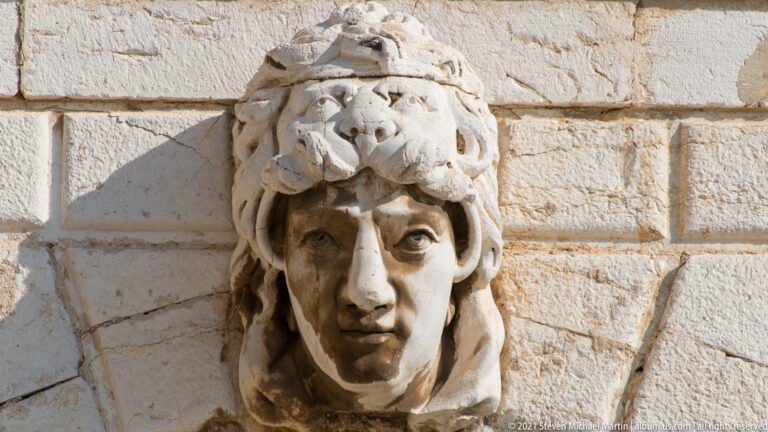 Carved stone human head projecting from a façade beneath an animal-pelt headdress with a muzzle above the forehead.