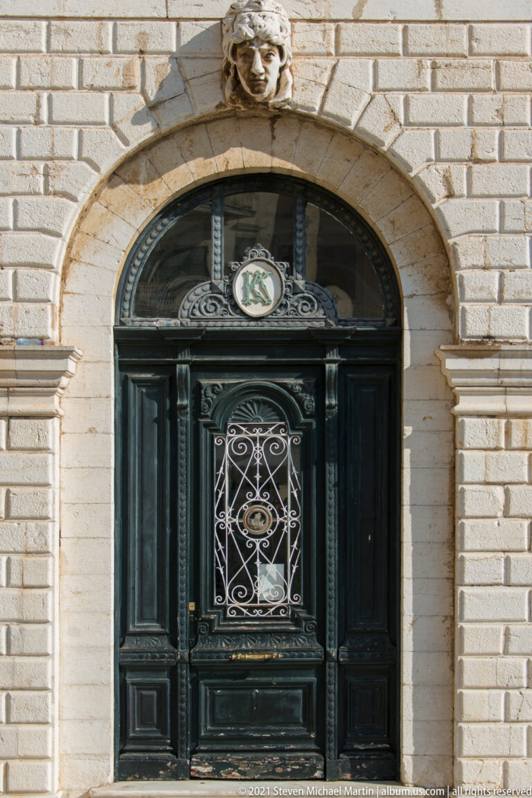 Ornate black arched doorway with paneled wood, scrolling wrought-iron grille, fanlight emblem, and carved stone head above.