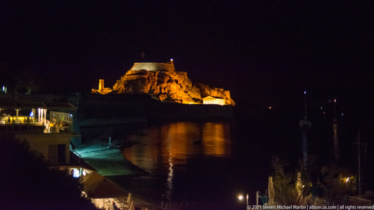 Floodlit Old Fortress of Corfu glowing above dark harbor water, with golden reflections below.