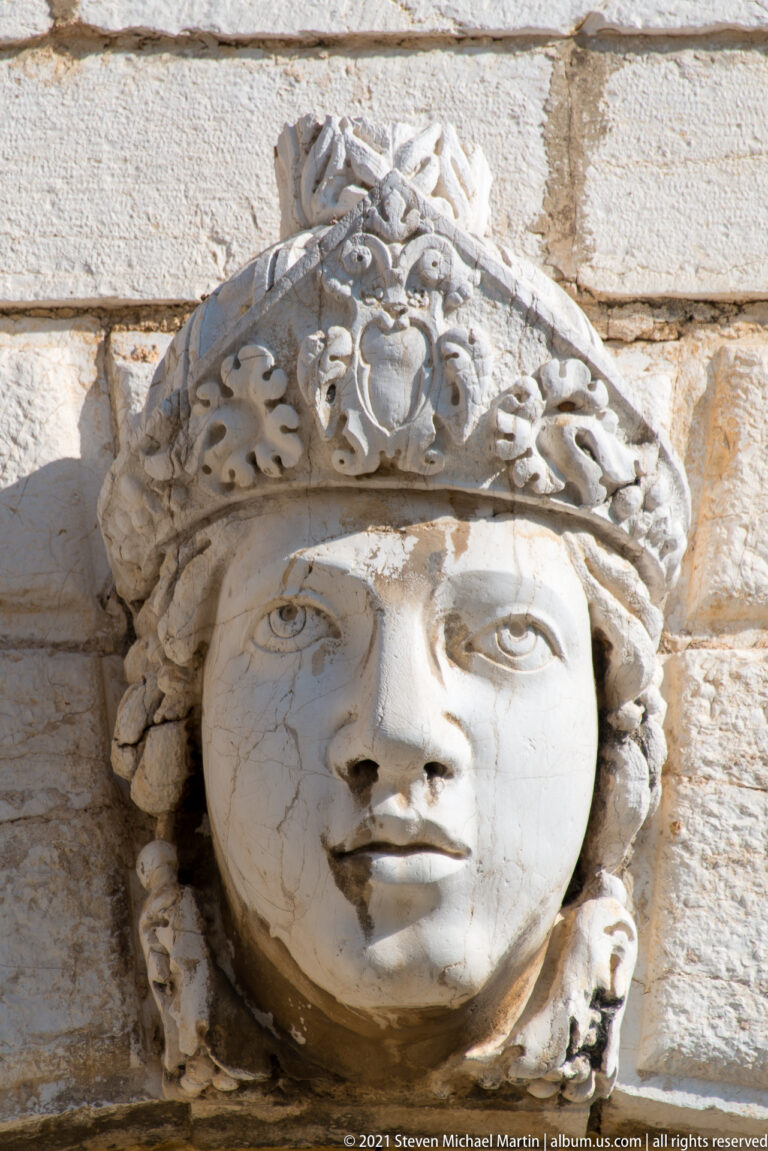 Close view of a pale stone female head with idealized features and an elaborate scrolling headdress projecting from a masonry wall.