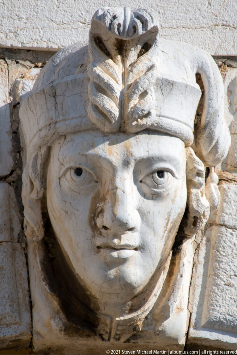 Close view of a pale stone female head with elaborate headdress projecting from a block-masonry façade.