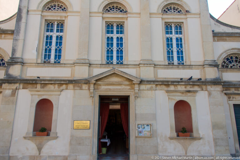 Church façade with central pedimented doorway, tall arched windows, pale pilasters, and open entrance.