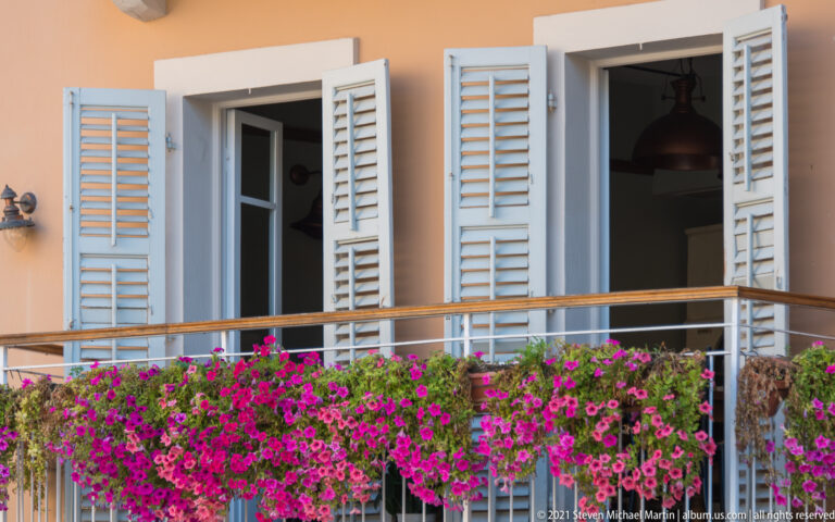 Peach façade with open white louvered shutters and a balcony densely planted with bright magenta flowers.
