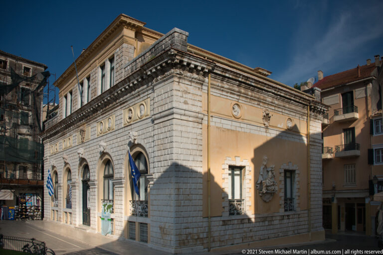 Historic Corfu corner building with rusticated stone base, ocher upper walls, arched openings, relief medallions, and Greek flag.