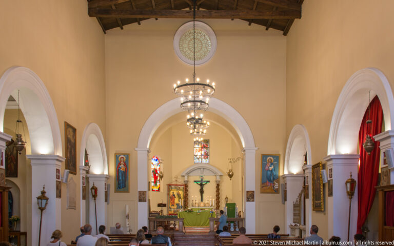 Church nave with white arches, painted icons, stained glass, tiered chandelier, timber ceiling, and seated worshippers.