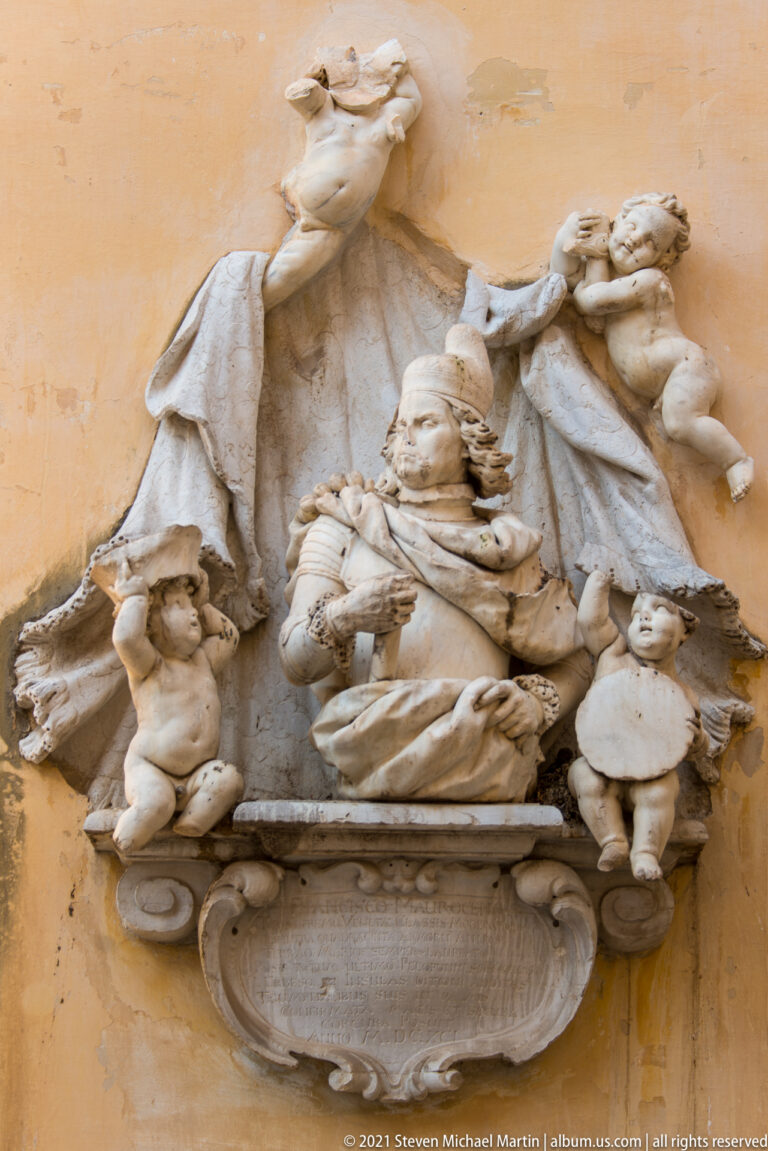 Elaborate pale stone wall relief with robed and winged figures arranged above a curved inscription tablet.