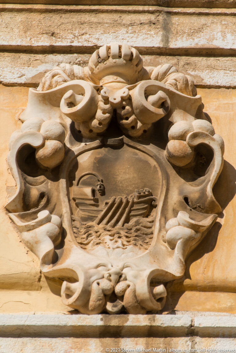 Weathered stone cartouche with central shield bearing a sailing vessel, surrounded by scrolls, shells, and floral ornament.