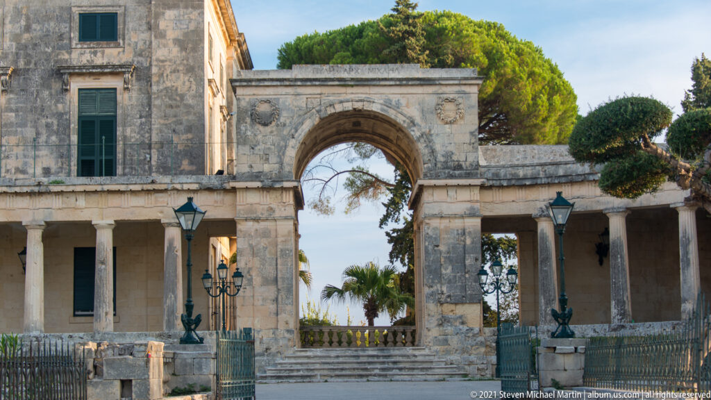 Stone entrance arch and colonnades of the Palace of Saints Michael and George, with green shutters, lamps, and garden trees.