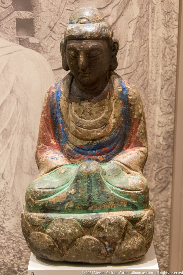 Worn polychrome seated Buddhist figure with hands folded in the lap and traces of red, blue, green, and gold pigment.