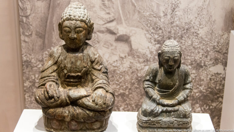 Two seated Buddha sculptures of different sizes, the larger weathered and pale, the smaller darker with more defined robe folds.