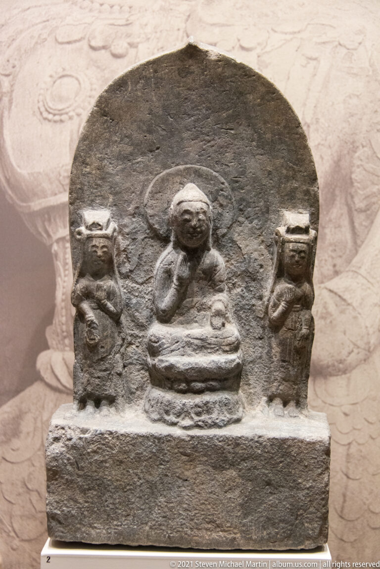 Dark stone Buddhist relief with a haloed seated Buddha at center and two smaller standing attendants on either side.