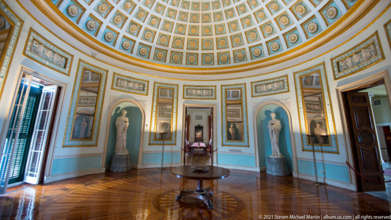Circular Rotunda with coffered dome, pale blue walls, mirrored panels, statue-filled niches, central table, and radial parquet floor.
