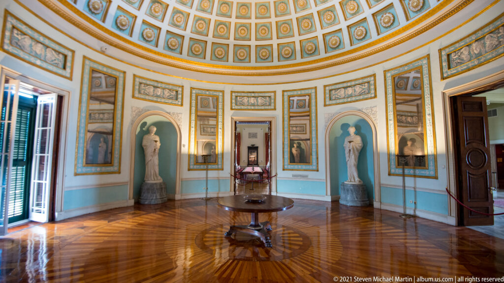 Wide view of the Rotunda with coffered dome, mirrored wall panels, statue niches, open shutters, central table, and radial parquet floor.