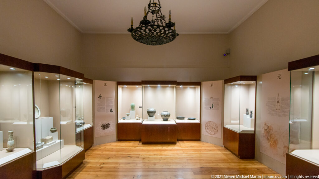 Museum gallery with wooden floor, illuminated glass display cases holding Asian ceramics and small objects, and a dark hanging lantern.