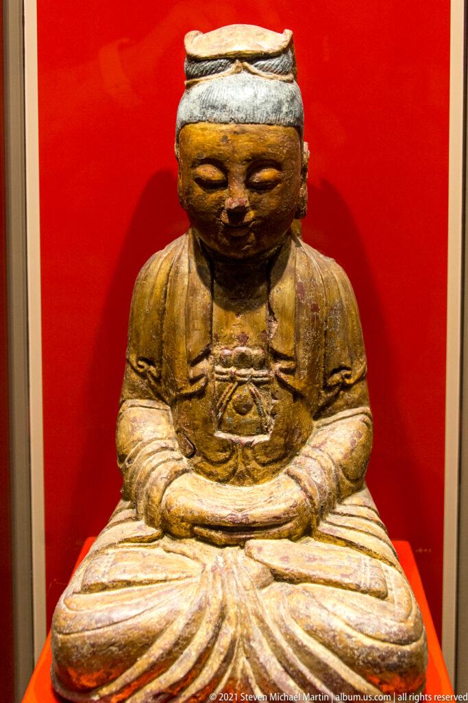 Worn painted seated religious figure with folded hands, layered robes, flat-topped head covering, and traces of ochre and red pigment.