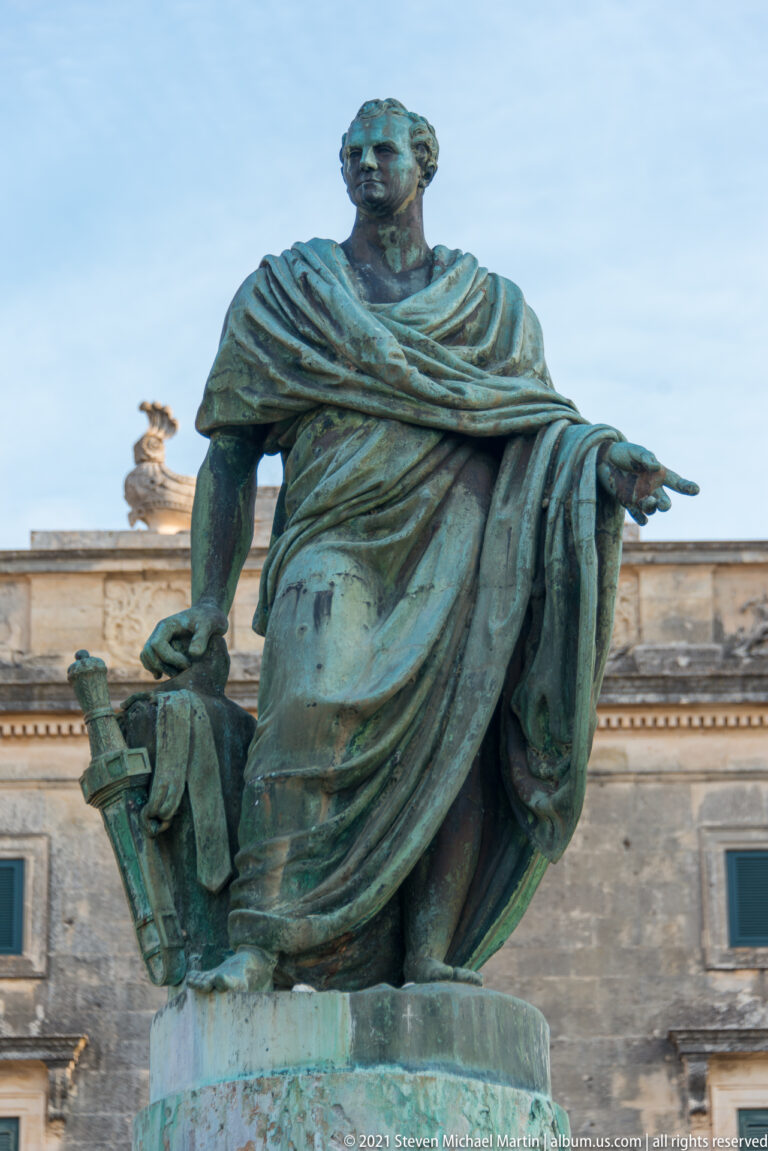 Green-patinated seated bronze statue of Sir Frederick Adam in classical drapery, with the pale stone palace behind him.