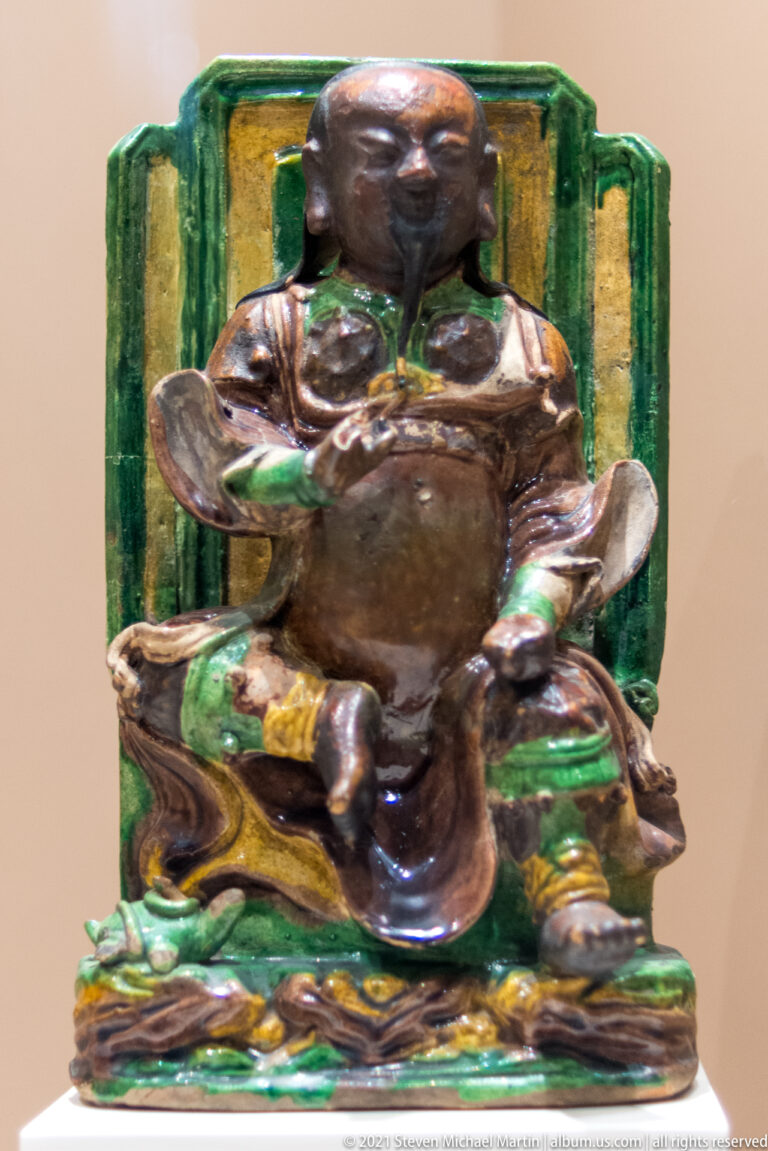 Brown-and-green glazed Chinese seated warrior or guardian figure with broad sleeves, heavy costume, and tall green backrest.