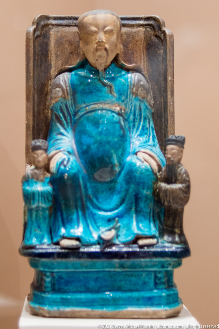 Turquoise-glazed seated Chinese male figure on a chair-like seat, with brown face and hands and a small vessel at his side.