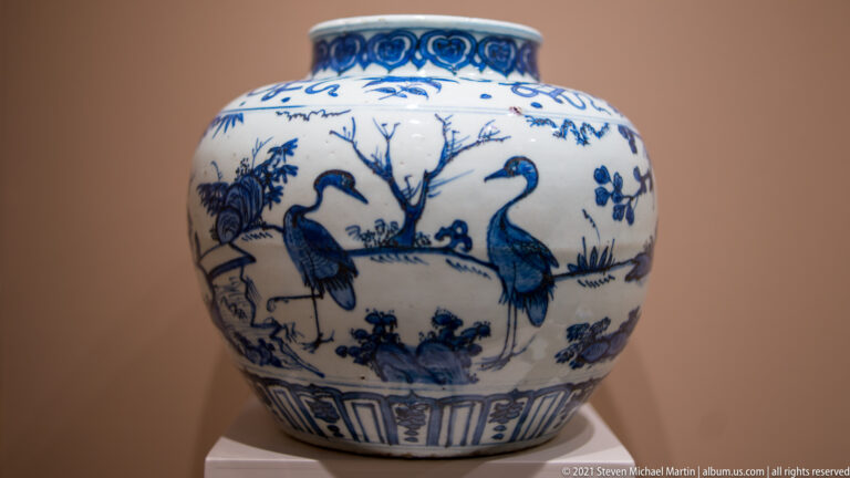 Blue-and-white porcelain jar with cobalt-painted long-legged birds, rocks, plants, and a bare tree beneath a decorated neck band.