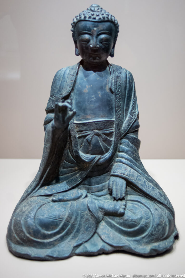 Dark bronze seated Buddha with curled hair, elongated ears, layered robe, right hand raised, and left hand resting near the knee.