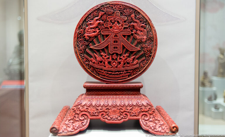 Circular red carved-lacquer Chinese plaque with dense figures and scrollwork, mounted on an ornate red pedestal stand.