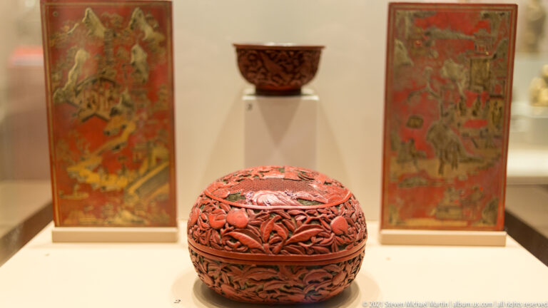 Round red carved-lacquer lidded box with dense floral scroll relief, displayed before two painted panels and a small cup.