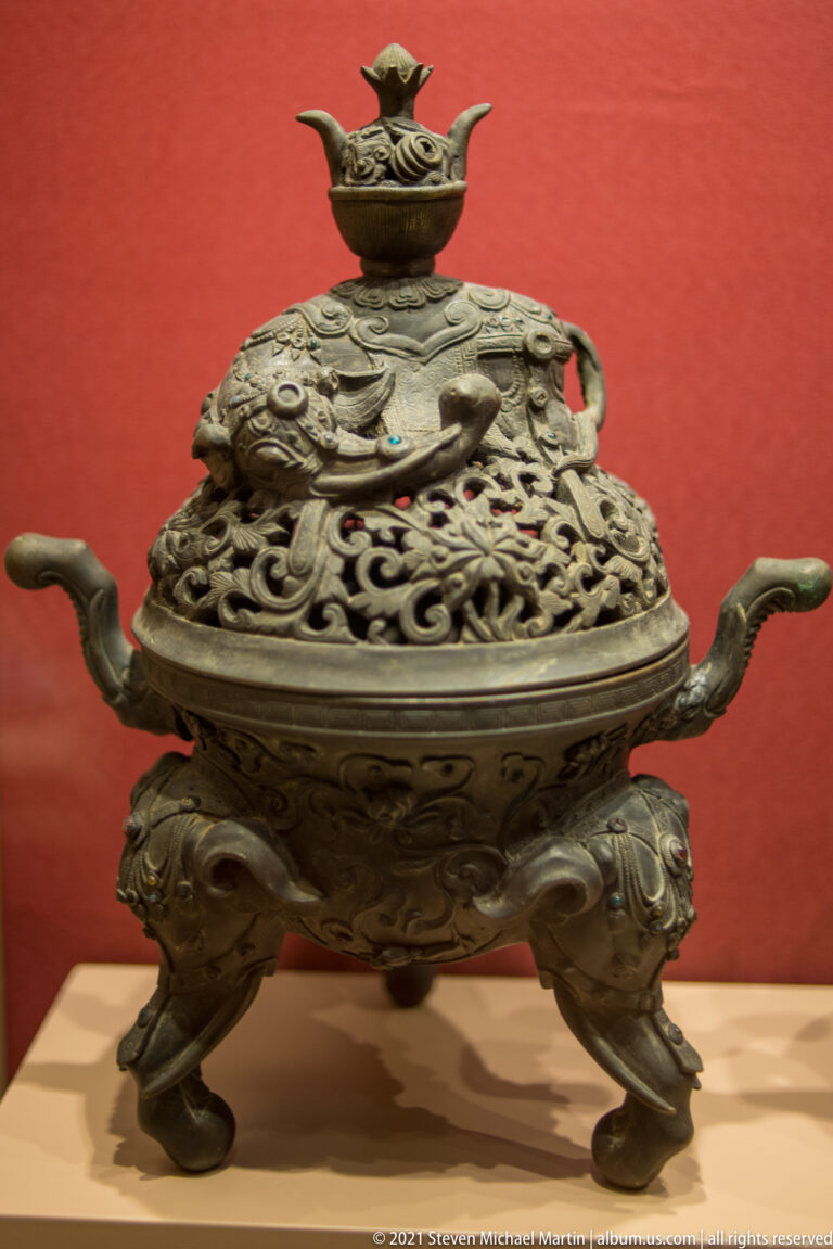 Ornate bronze tripod incense burner with curved handles, pierced domed lid, scrolling relief, and a small bird finial.
